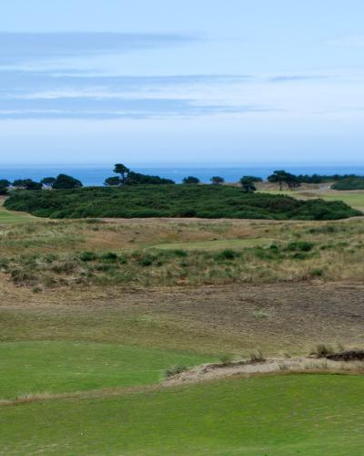 A landscape view of a golf course with rolling green fairways, sand bunkers, and a line of trees in the distance, with the ocean visible on the horizon.