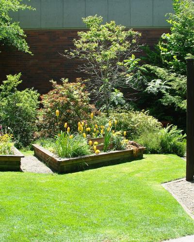 The image shows a landscaped garden area with raised wooden planters, green grass, and a prominent dark wooden sign on the right that reads "Martha Spring Botanical Garden" (or similar, partially obscured but clearly a garden entrance sign).