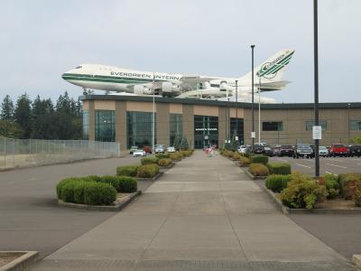 The image shows the exterior of a building with a large, white Boeing 747-400 aircraft mounted on its roof; the building's facade has a sign that reads "WINGS & WAVE" and the aircraft has "EVERGREEN" written on its fuselage.