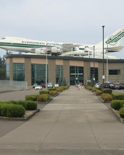 The image shows the exterior of a building with a large, white Boeing 747-400 aircraft mounted on its roof; the building's facade has a sign that reads "WINGS & WAVE" and the aircraft has "EVERGREEN" written on its fuselage.