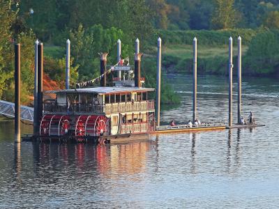 A sternwheel paddlewheel boat (the Willamette Queen) moored at a dock, with a metal gangway, green riverbank, and a person in a kayak nearby.