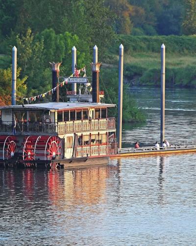 A sternwheel paddlewheel boat (the Willamette Queen) moored at a dock, with a metal gangway, green riverbank, and a person in a kayak nearby.