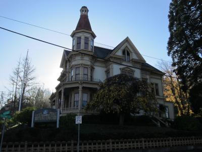 A large, ornate Victorian-style house with a prominent corner turret and cupola, viewed from a street level with a sign in the foreground.
