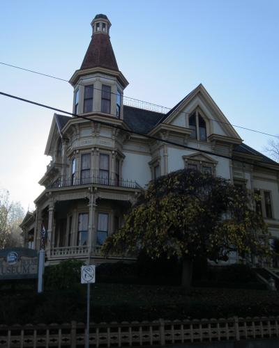 A large, ornate Victorian-style house with a prominent corner turret and cupola, viewed from a street level with a sign in the foreground.