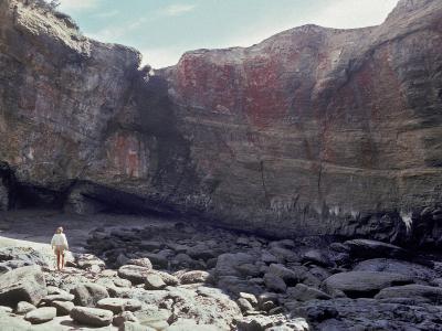 The image shows a recess in a rocky cliff face, likely a sea cave or cove, with a person standing on the rocky shore in the foreground; the rock face is layered and features a distinct reddish-brown stain on the upper section.