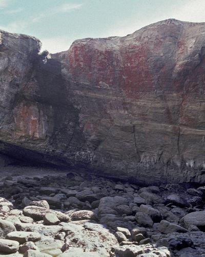 The image shows a recess in a rocky cliff face, likely a sea cave or cove, with a person standing on the rocky shore in the foreground; the rock face is layered and features a distinct reddish-brown stain on the upper section.