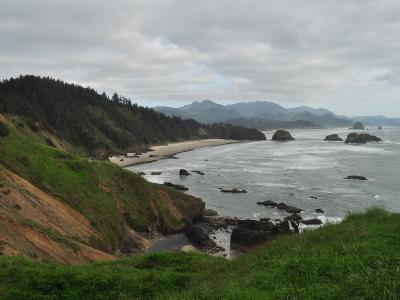 A coastal landscape view showing a sandy beach (Trestle Cove/Crescent Beach) below a grassy, forested bluff, with the ocean and sea stacks (Haystack Rock and others) in the distance.