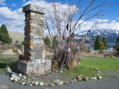 A stone monument with a plaque, situated in a grassy area, with a tree to its right and a body of water and mountains in the background.