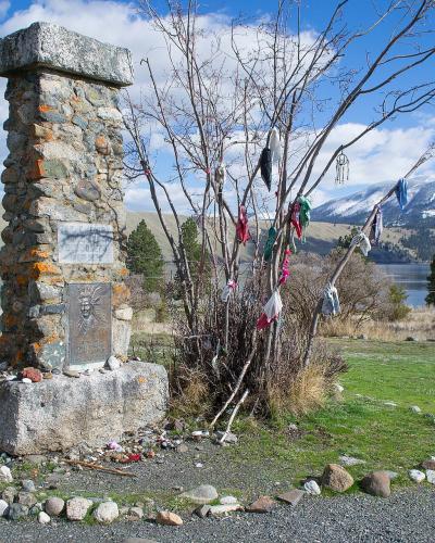 A stone monument with a plaque, situated in a grassy area, with a tree to its right and a body of water and mountains in the background.