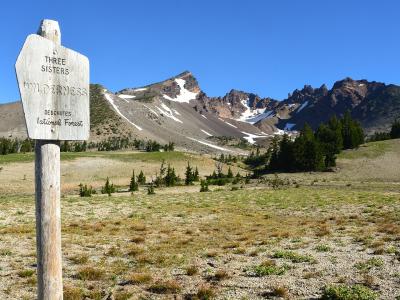A wooden sign reading "THREE SISTER WILDERNESS" stands in the foreground, with a view of rugged, snow-dusted mountain peaks (the Three Sisters) in the background.