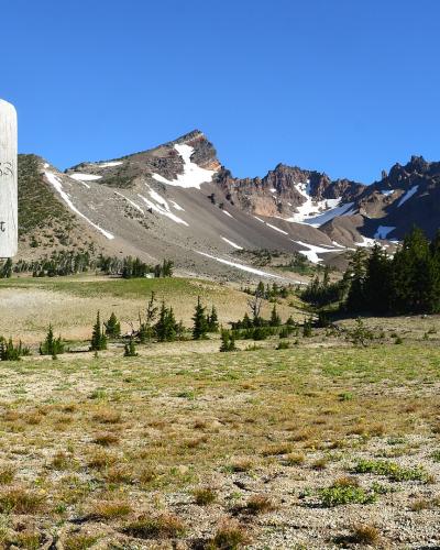 A wooden sign reading "THREE SISTER WILDERNESS" stands in the foreground, with a view of rugged, snow-dusted mountain peaks (the Three Sisters) in the background.