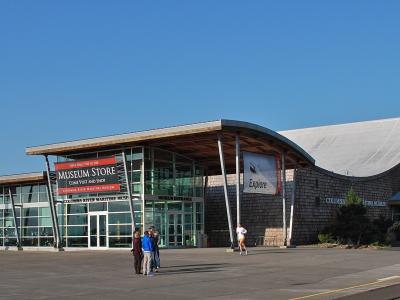 The image shows the exterior of a modern building with a distinctive curved roof and shingle siding, featuring a large red banner that reads "MUSEUM STORE" and text on the building facade identifying it as the "COLUMBIA RIVER MARITIME MUSEUM".