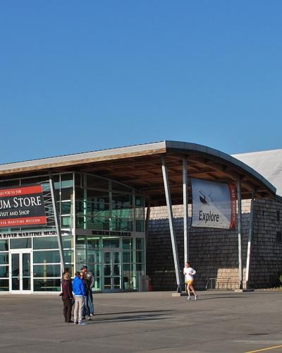 The image shows the exterior of a modern building with a distinctive curved roof and shingle siding, featuring a large red banner that reads "MUSEUM STORE" and text on the building facade identifying it as the "COLUMBIA RIVER MARITIME MUSEUM".