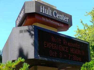The image shows a large outdoor marquee sign for the "Hult Center for the Performing Arts" with an electronic display board underneath showing event information against a blue sky.