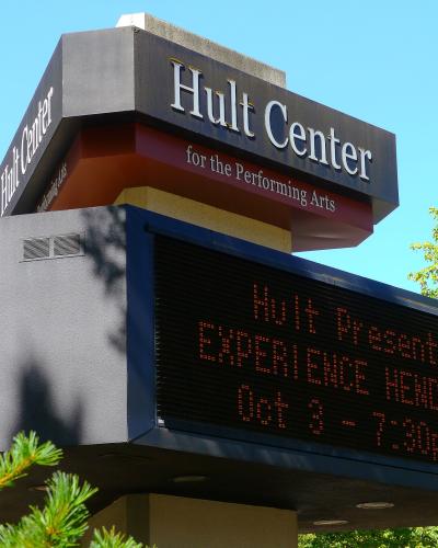 The image shows a large outdoor marquee sign for the "Hult Center for the Performing Arts" with an electronic display board underneath showing event information against a blue sky.