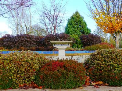 A stone monument or planter with the text "MADD GARDEN" on a plaque, surrounded by colorful bushes and trees.