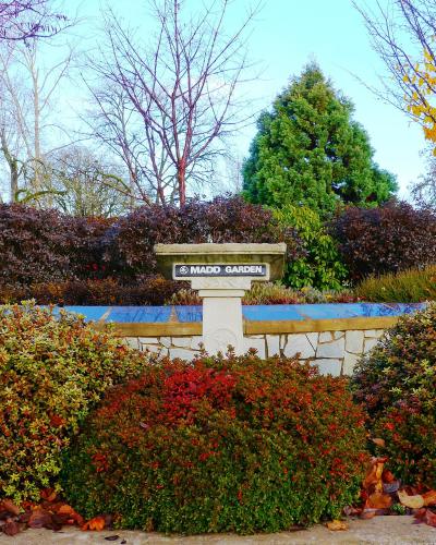 A stone monument or planter with the text "MADD GARDEN" on a plaque, surrounded by colorful bushes and trees.