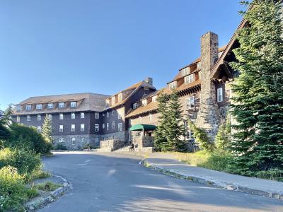 A large, multi-story building with a rustic, lodge-style architecture, featuring a prominent stone chimney, brown shingled walls, and white-framed windows, set against a clear blue sky.
