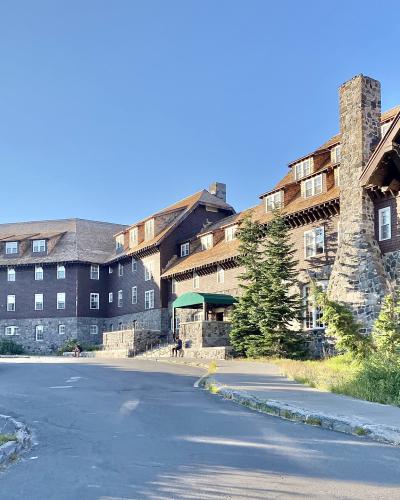 A large, multi-story building with a rustic, lodge-style architecture, featuring a prominent stone chimney, brown shingled walls, and white-framed windows, set against a clear blue sky.