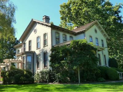A large, multi-story, light-colored Victorian-style house with a complex roofline, surrounded by mature trees and green lawn, with a white pergola structure visible to the left.