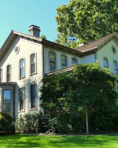 A large, multi-story, light-colored Victorian-style house with a complex roofline, surrounded by mature trees and green lawn, with a white pergola structure visible to the left.