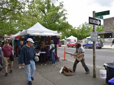 A street scene showing a street musician playing a guitar on a sidewalk, with white vendor tents set up behind him and a street sign for "10th St" and "F St" visible on a pole.