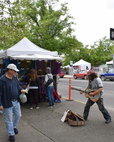 A street scene showing a street musician playing a guitar on a sidewalk, with white vendor tents set up behind him and a street sign for "10th St" and "F St" visible on a pole.