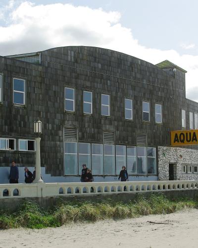 The image shows a large, dark-shingled building with a prominent yellow sign reading "AQUARIUM" on its side, situated on a sandy beach with people walking nearby.