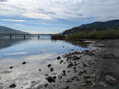 A view of a rocky shoreline and calm water with a long truss bridge spanning the river in the distance, flanked by hills.
