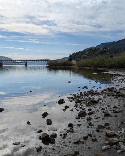 A view of a rocky shoreline and calm water with a long truss bridge spanning the river in the distance, flanked by hills.