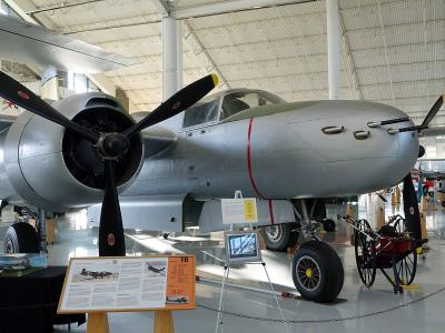 A silver Douglas A-26C Invader aircraft is displayed indoors on a polished floor within a large hangar-like museum space, with informational placards and other aircraft visible in the background.