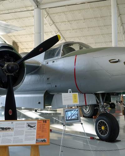 A silver Douglas A-26C Invader aircraft is displayed indoors on a polished floor within a large hangar-like museum space, with informational placards and other aircraft visible in the background.