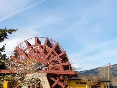 A large red wooden water wheel stands on a concrete base in a grassy yard, with a single-story yellow building behind it that has the word "MUSEUM" painted in black capital letters on its facade.