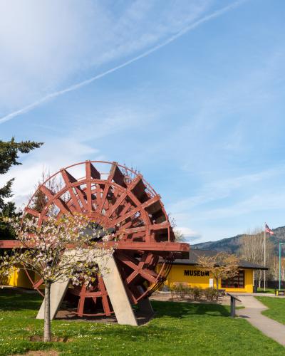 A large red wooden water wheel stands on a concrete base in a grassy yard, with a single-story yellow building behind it that has the word "MUSEUM" painted in black capital letters on its facade.