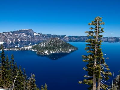 A scenic landscape photograph showing Crater Lake in Oregon, featuring Wizard Island rising from the deep blue water, surrounded by snow-dusted caldera walls and evergreen trees in the foreground.