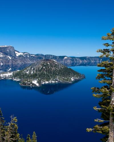 A scenic landscape photograph showing Crater Lake in Oregon, featuring Wizard Island rising from the deep blue water, surrounded by snow-dusted caldera walls and evergreen trees in the foreground.