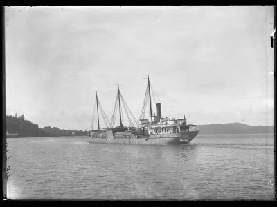 A black and white photograph showing a three-masted steamship with a single smokestack moving through water, with a forested shoreline visible in the background.