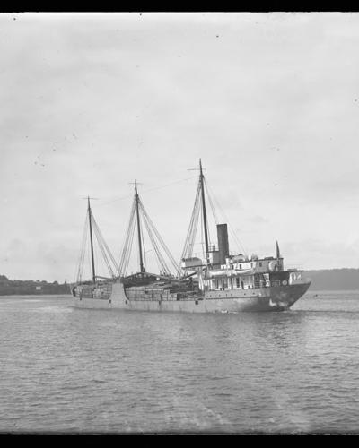A black and white photograph showing a three-masted steamship with a single smokestack moving through water, with a forested shoreline visible in the background.