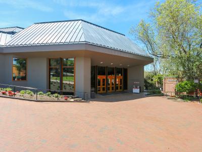 The image shows the exterior entrance of a modern building with a distinctive metal roof, large glass windows, and orange-framed doors, situated on a brick plaza with landscaping.