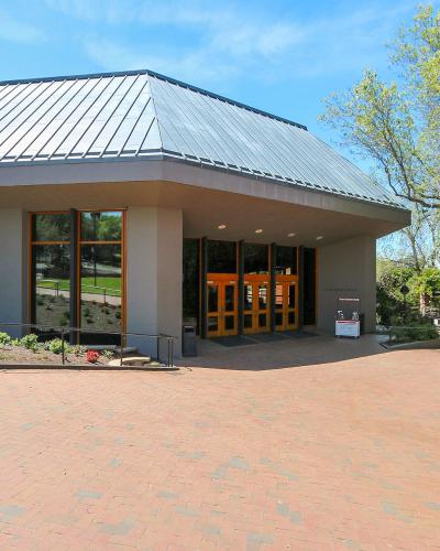 The image shows the exterior entrance of a modern building with a distinctive metal roof, large glass windows, and orange-framed doors, situated on a brick plaza with landscaping.