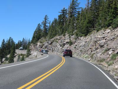 A view from a moving vehicle looking down a two-lane paved road winding through a forest of coniferous trees, with a mountain slope visible in the background.