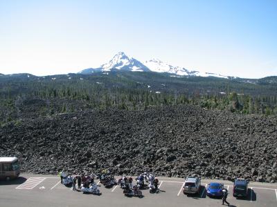 A view from a paved parking lot looking out over a dark, solidified lava field towards a distant, snow-capped stratovolcano (Mount Hood) under a clear sky, with a group of motorcyclists gathered in the foreground.