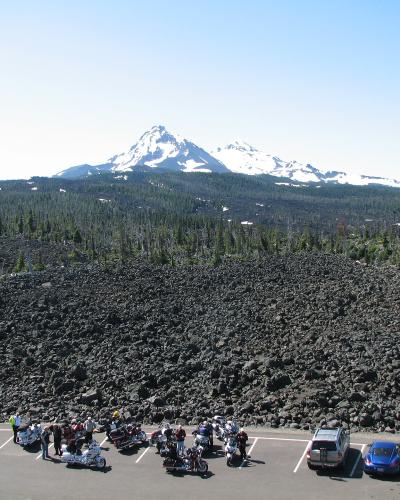 A view from a paved parking lot looking out over a dark, solidified lava field towards a distant, snow-capped stratovolcano (Mount Hood) under a clear sky, with a group of motorcyclists gathered in the foreground.