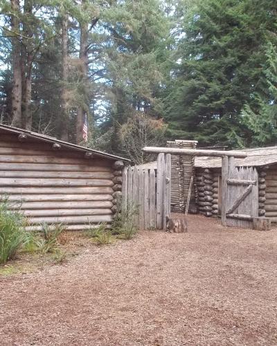 A reconstruction of a historic log fort with wooden palisades, a gate, and an American flag, set within a forest of tall evergreen trees.