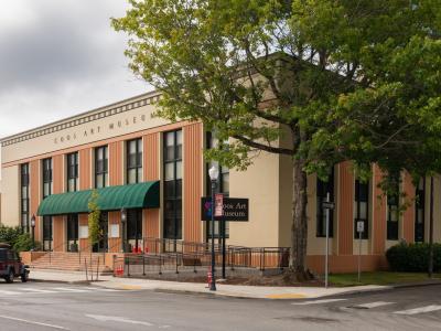 A beige and terracotta-colored building with vertical architectural accents, a green awning over the entrance, and large lettering on the facade reading "COOS ART MUSEUM".