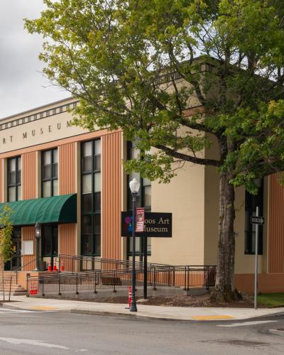 A beige and terracotta-colored building with vertical architectural accents, a green awning over the entrance, and large lettering on the facade reading "COOS ART MUSEUM".