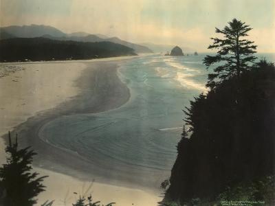 A vintage colorized photograph showing a wide sandy beach, ocean waves, and sea stacks in the distance, viewed from an elevated cliffside position with trees in the foreground.