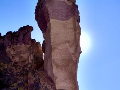 A low-angle shot of a tall, slender, light-colored rock spire (a pinnacle) against a blue sky, with a small figure of a rock climber visible on the rock face.