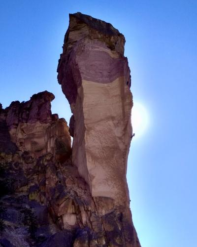 A low-angle shot of a tall, slender, light-colored rock spire (a pinnacle) against a blue sky, with a small figure of a rock climber visible on the rock face.