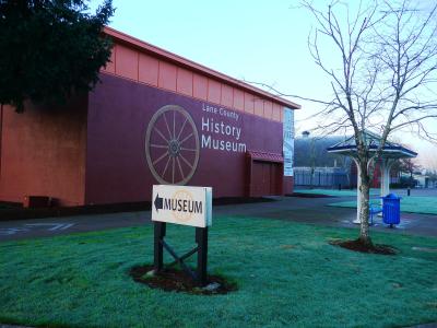 The image shows the exterior of a building with a large, painted wooden wagon wheel on its reddish-brown wall, a white sign in the foreground reading "MUSEUM" with an arrow, and a blue trash can on the grass.